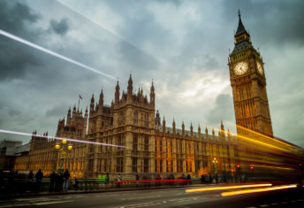Big Ben and the Houses of Parliament in London, England. The building is lit up and set against a dramatic sky with cars and busses zooming by on Waterloo Bridge.