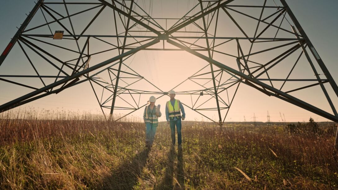 Male and Female Technicians Walking Under Power Transmission Tower in Dry Grass Field During Warm Sunlight Getty, Male and female technicians walking through dry grass field under power transmission tower. Wearing safety helmets and vest while moving forward. Warm sunlight creating strong industrial and energy scene.